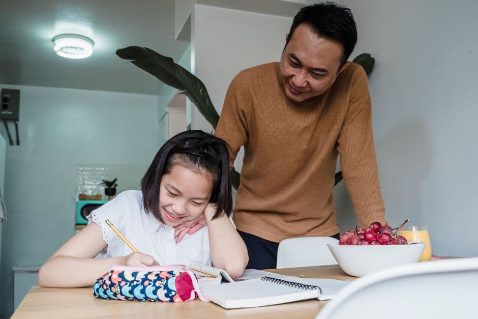 A primary-school child working through homework at a tidy kitchen table while a parent sits calmly nearby.
