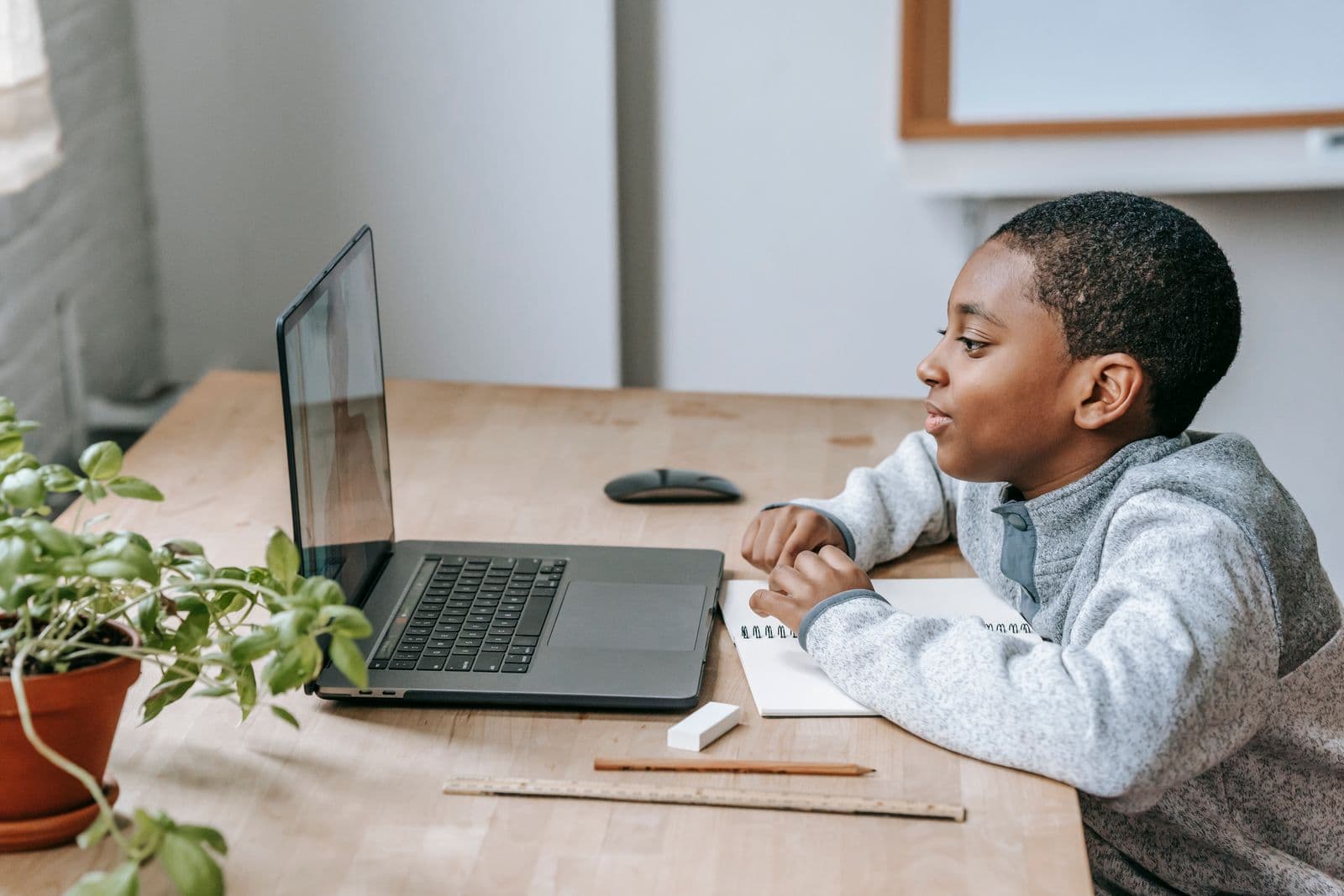A calm primary-school boy working at a laptop with a notebook beside him.