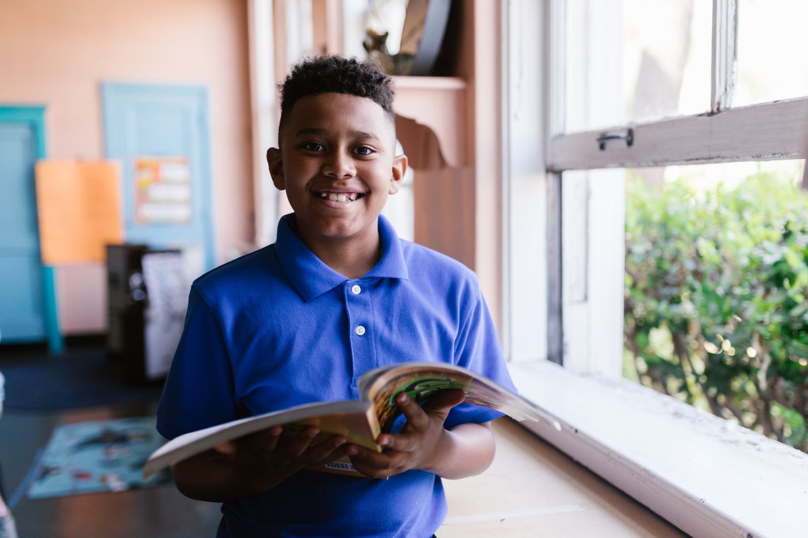 A smiling primary-school boy holding an open book by a sunlit window.