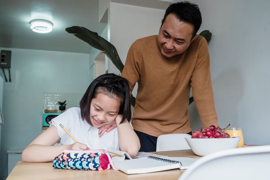A primary-school child working through homework at a tidy kitchen table while a parent sits calmly nearby.