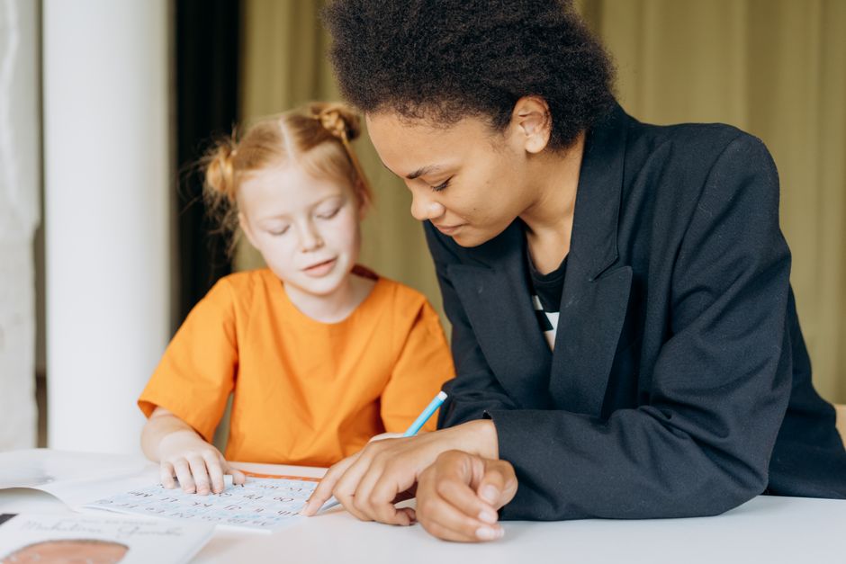 A primary-school child working through a Maths worksheet on paper beside an educator at a learning centre table.